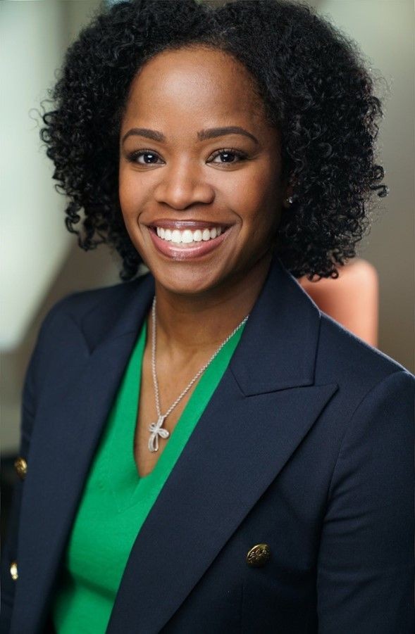 Smiling woman with curly hair wearing a navy blazer and green top, sitting indoors.